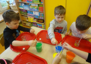 Troje dzieci nabiera zabarwioną ciesz stojącą w pojemnikach na środku stolika.
